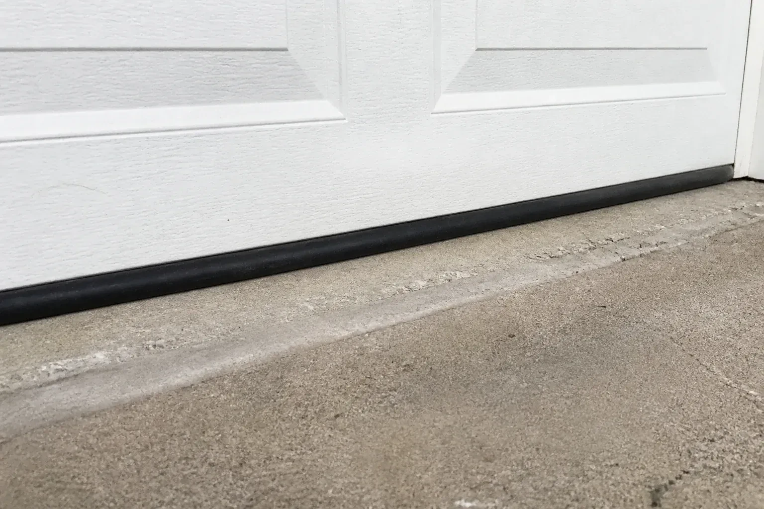 Black bottom seal along a closed white garage door at a concrete threshold. — Garage Door Bottom Seal Replacement, West Grey